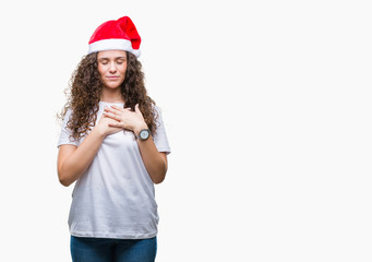 Young brunette girl wearing christmas hat over isolated background smiling with hands on chest with closed eyes and grateful gesture on face. Health concept.