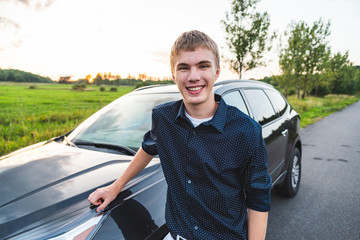 Happy young man leaning against his car next to an open field at sunset.