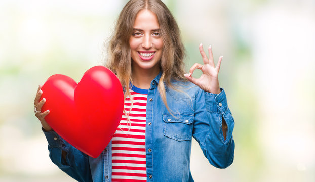 Beautiful young blonde woman holding heart valentine over isolated background doing ok sign with fingers, excellent symbol