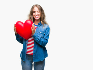 Beautiful young blonde woman holding heart valentine over isolated background with a happy face standing and smiling with a confident smile showing teeth