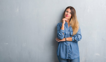 Fototapeta premium Young adult woman over grunge grey wall wearing denim outfit with hand on chin thinking about question, pensive expression. Smiling with thoughtful face. Doubt concept.