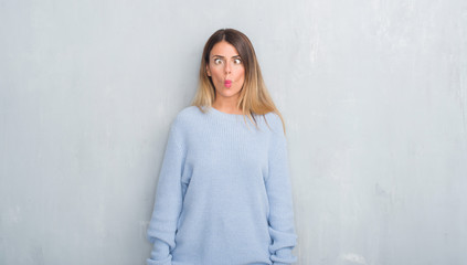 Young adult woman over grey grunge wall wearing winter outfit making fish face with lips, crazy and comical gesture. Funny expression.