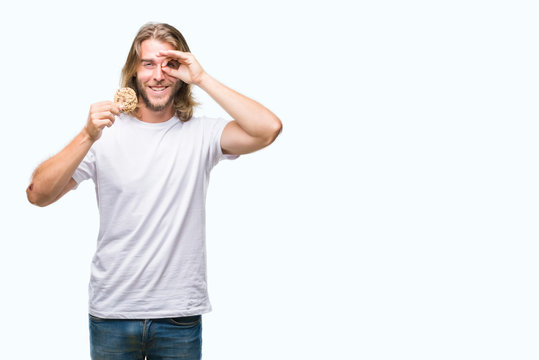 Young handsome man with long hair eating chocolate cooky over isolated background with happy face smiling doing ok sign with hand on eye looking through fingers