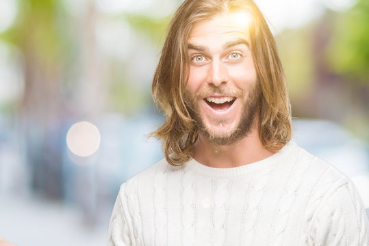 Young handsome man with long hair wearing winter sweater over isolated background amazed and smiling to the camera while presenting with hand and pointing with finger.