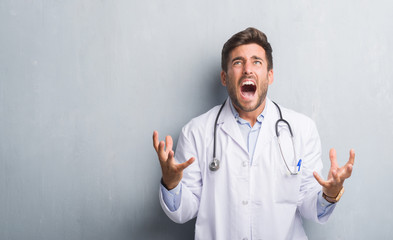 Handsome young doctor man over grey grunge wall crazy and mad shouting and yelling with aggressive expression and arms raised. Frustration concept.