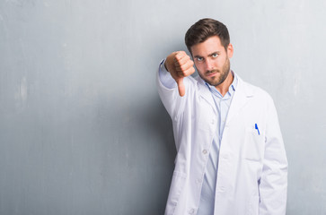 Handsome young professional man over grey grunge wall wearing white coat looking unhappy and angry showing rejection and negative with thumbs down gesture. Bad expression.