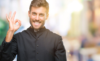 Young catholic christian priest man over isolated background smiling positive doing ok sign with hand and fingers. Successful expression.