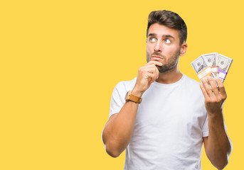 Young handsome man holding stack of dollars over isolated background serious face thinking about...