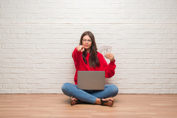 Young brunette woman sitting on the floor over white brick wall paying holding dollars pointing with finger to the camera and to you, hand sign, positive and confident gesture from the front