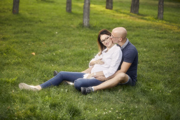Naklejka na ściany i meble young couple sitting in grass field in park. smiling happy, man kissing pregnant woman.