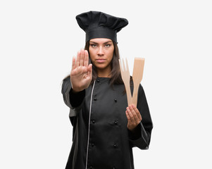 Young hispanic cook woman wearing chef uniform with open hand doing stop sign with serious and confident expression, defense gesture