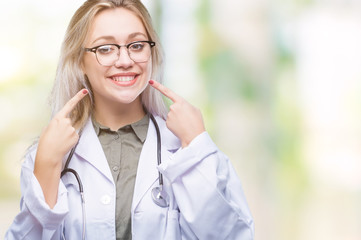 Young blonde doctor woman over isolated background smiling confident showing and pointing with fingers teeth and mouth. Health concept.