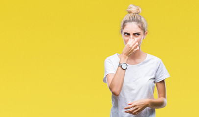 Young beautiful blonde woman wearing white t-shirt over isolated background smelling something stinky and disgusting, intolerable smell, holding breath with fingers on nose. Bad smells concept.