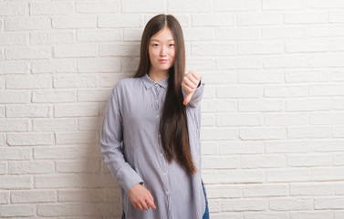 Young Chinese woman over brick wall with angry face, negative sign showing dislike with thumbs...