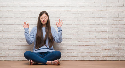 Young Chinese woman sitting on the floor over brick wall relax and smiling with eyes closed doing meditation gesture with fingers. Yoga concept.