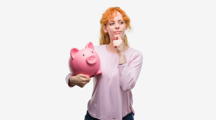 Young redhead woman holding piggy bank serious face thinking about question, very confused idea
