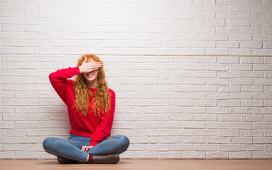 Young redhead woman sitting over brick wall smiling and laughing with hand on face covering eyes for surprise. Blind concept.