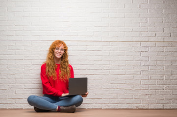 Young redhead woman sitting over brick wall using computer laptop with a happy face standing and smiling with a confident smile showing teeth