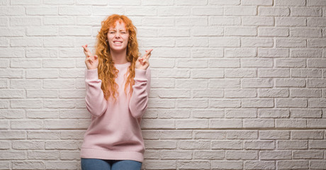 Young redhead woman standing over brick wall smiling crossing fingers with hope and eyes closed. Luck and superstitious concept.