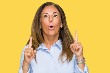 Beautiful middle age business adult woman over isolated background amazed and surprised looking up and pointing with fingers and raised arms.