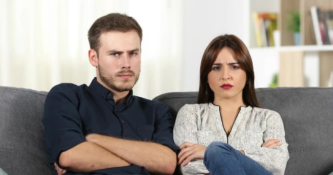 Front view of an angry couple looking at camera sitting on a couch at home