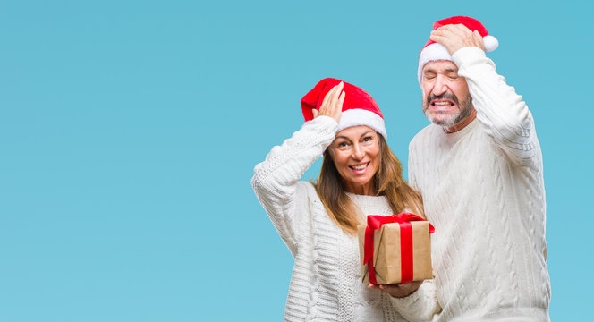 Middle age hispanic couple wearing christmas hat and holding gift over isolated background stressed with hand on head, shocked with shame and surprise face, angry and frustrated. Fear and upset