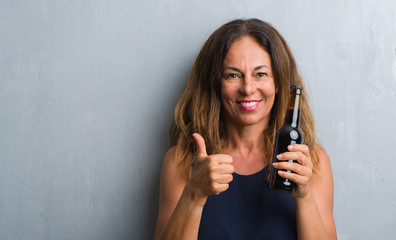 Middle age hispanic woman standing over grey grunge wall holding beer bottle happy with big smile doing ok sign, thumb up with fingers, excellent sign