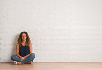 Middle age hispanic woman sitting on the floor over white brick wall puffing cheeks with funny face. Mouth inflated with air, crazy expression.