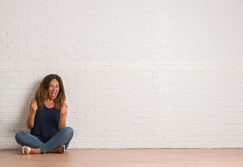 Middle age hispanic woman sitting on the floor over white brick wall very happy and excited doing winner gesture with arms raised, smiling and screaming for success. Celebration concept.