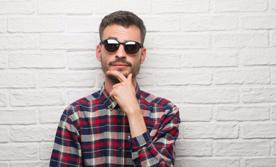 Young adult man wearing sunglasses standing over white brick wall serious face thinking about...