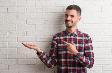 Young adult man standing over white brick wall amazed and smiling to the camera while presenting with hand and pointing with finger.