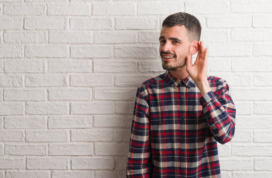 Young adult man standing over white brick wall smiling with hand over ear listening an hearing to rumor or gossip. Deafness concept.