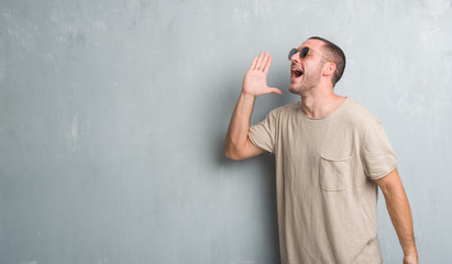 Young caucasian man over grey grunge wall wearing sunglasses shouting and screaming loud to side with hand on mouth. Communication concept.