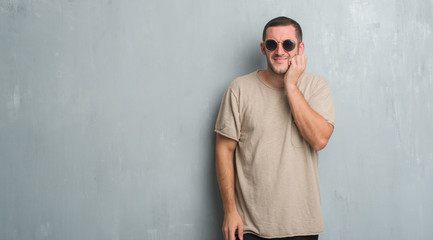 Young caucasian man over grey grunge wall wearing sunglasses touching mouth with hand with painful expression because of toothache or dental illness on teeth. Dentist concept.