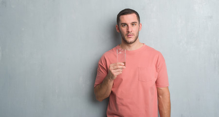 Young caucasian man over grey grunge wall drinking a glass of water with a confident expression on smart face thinking serious