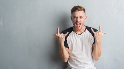 Young redhead man over grey grunge wall shouting with crazy expression doing rock symbol with hands...