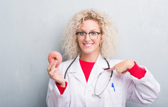 Young blonde dietist doctor woman over grunge grey wall holding donut with surprise face pointing finger to himself