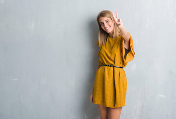 Naklejka premium Beautiful young woman standing over grunge grey wall wearing a dress smiling looking to the camera showing fingers doing victory sign. Number two.