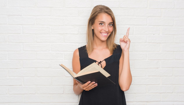 Beautiful young woman over white brick wall reading a book surprised with an idea or question pointing finger with happy face, number one