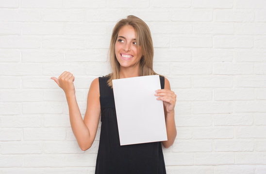 Beautiful young woman over white brick wall holding blank paper sheet pointing and showing with thumb up to the side with happy face smiling