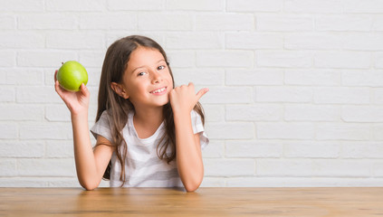 Young hispanic kid sitting on the table eating fresh green apple pointing and showing with thumb up to the side with happy face smiling
