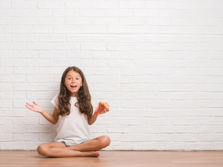 Young hispanic kid sitting on the floor over white brick wall eating pizza slice very happy and...