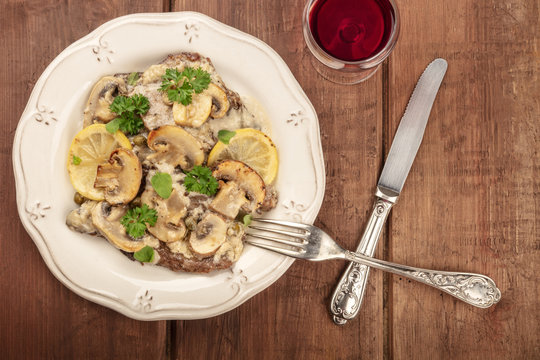 A photo of scallopine di vitello, veal scallopini, with a mushroom sauce, lemons, and parsley, shot from above on a dark wooden texture with red wine and copyspace