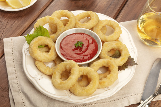 A closeup photo of squid rings with a tomato sauce, lemon wedges, and white wine, on a dark rustic wooden background