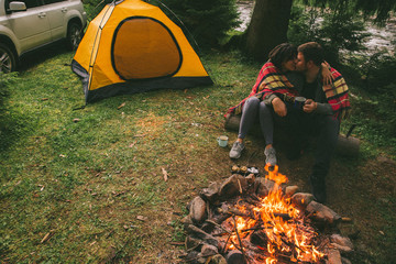 couple sitting near camp fire and drinking tea and telling stories . tent and suv on background