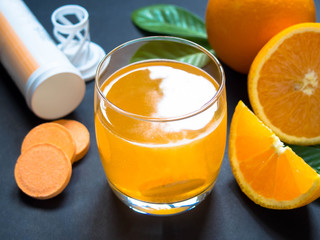 Vitamin c effervescent tablet is dissolving in glass of water w/ medicine bottle and fresh juicy orange fruits on dark table. Concept of vitamin/mineral supplement, nutrition, health care and medical.