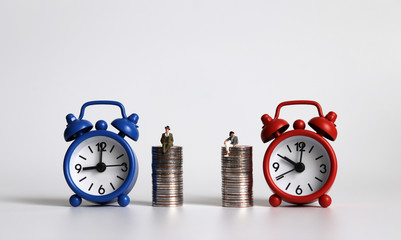 Miniature man and miniature woman sitting on a pile of coins at different alarm clocks.