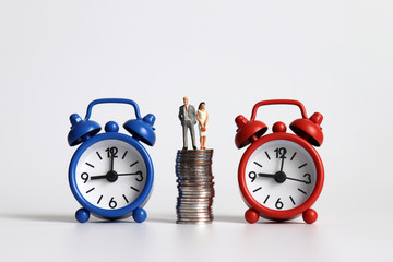 Miniature man and miniature woman standing on a pile of coins between two alarm clocks.