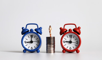 Miniature businesswoman standing on a pile of coins between two alarm clocks.