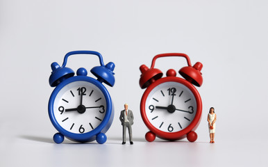 Miniature man standing next to the blue alarm clock and miniature woman standing next to the red alarm clock.